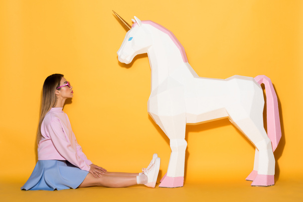 side view of asian female model in sunglasses sitting on floor and decorative unicorn on yellow background  - Fotografie, Obrázek