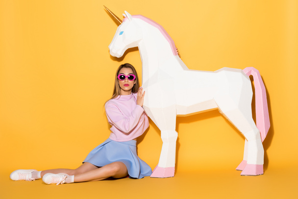 young female model in sunglasses sitting on floor and embracing decorative unicorn on yellow background - Foto, afbeelding