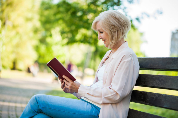 Adult woman sitting in park and reading book. - Фото, изображение