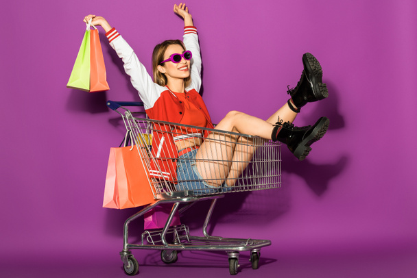 hermosa mujer joven feliz en gafas de sol sosteniendo bolsas de papel y sentado en el carro de la compra en violeta
  - Foto, Imagen