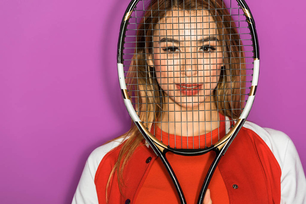 beautiful asian girl looking at camera through tennis racquet isolated on violet - Fotó, kép