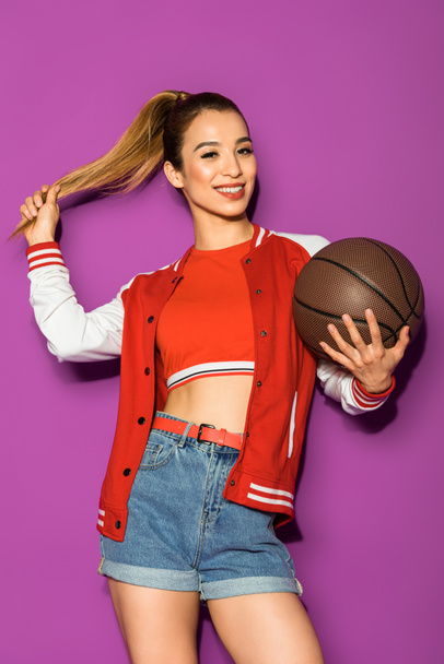 beautiful cheerful asian girl holding basketball ball and smiling at camera isolated on violet - Fotó, kép