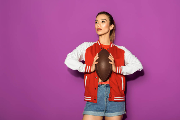 beautiful asian woman holding rugby ball and looking away isolated on violet - Fotoğraf, Görsel