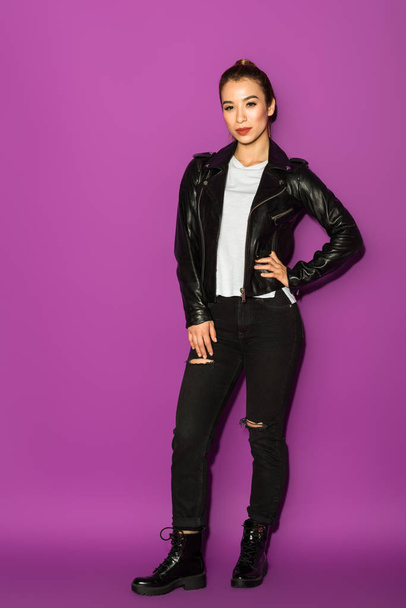 full length view of beautiful stylish asian girl in leather jacket looking at camera isolated on violet - Foto, immagini