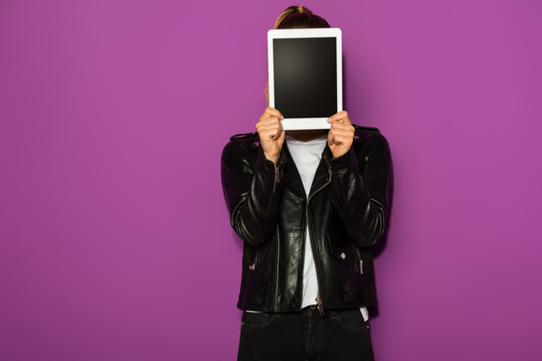 stylische junge Frau in Lederjacke versteckt Gesicht mit digitalem Tablet isoliert auf violett - Foto, Bild