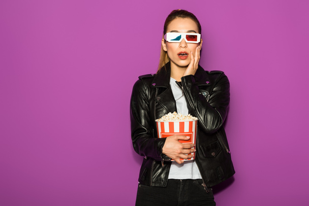 šokovaná mladá žena v 3d brýlích jíst popcorn a při pohledu na fotoaparát izolované na fialové  - Fotografie, Obrázek