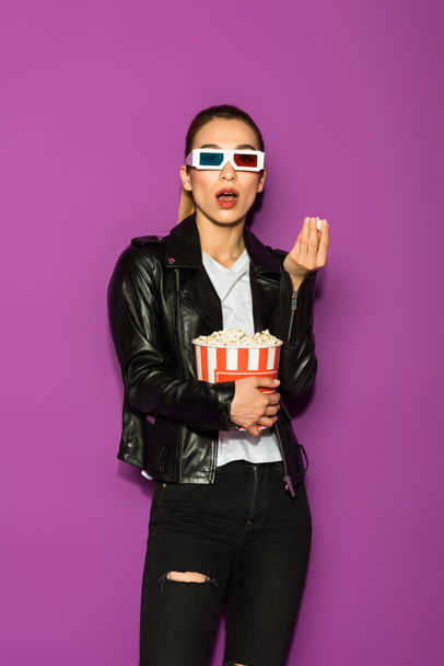 schockiertes asiatisches Mädchen in Lederjacke und 3D-Brille, das Popcorn isst und isoliert auf Veilchen in die Kamera schaut  - Foto, Bild