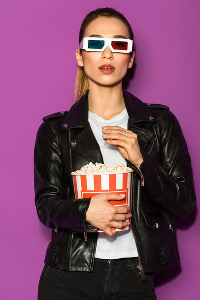 piękna dziewczyna azjatyckich w skórzanej kurtce i okulary 3d jedzenie popcorn i patrząc na kamery na białym tle na fioletowo - Zdjęcie, obraz
