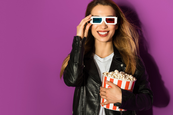 beautiful young woman in 3d glasses holding box of popcorn and smiling at camera isolated on violet - Photo, image