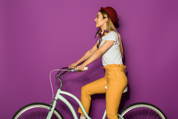 attractive smiling stylish girl sitting on bicycle and looking away isolated on violet - Fotografie, Obrázek
