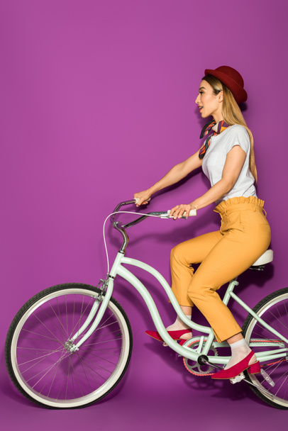 Widok z boku atrakcyjne stylowa dziewczyna azjatyckich jazdy na rowerze i patrząc od hotelu na białym tle na fioletowo - Zdjęcie, obraz