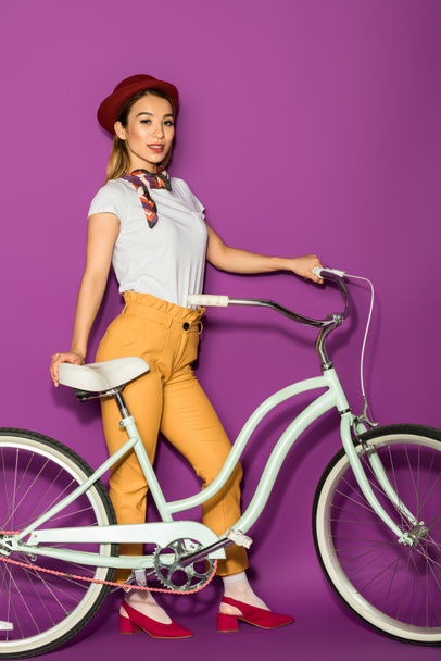 full length of beautiful stylish asian woman standing with bike and smiling at camera isolated on violet  - Foto, Imagem