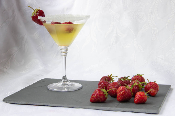 Ένα κοκτέιλ κίτρινο χρώμα σε ένα ποτήρι διακοσμημένο με φράουλες σε ένα ταμπλό με γκρίζα πέτρα με διάσπαρτα φράουλες - Φωτογραφία, εικόνα
