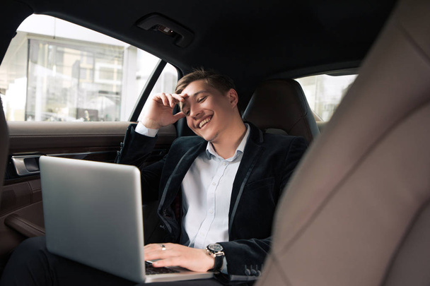 Seid immer fröhlich und positiv! fröhlich anerkannter junger professioneller Geschäftsmann sitzt im Auto und lächelt, während er am Laptop arbeitet. - Foto, Bild