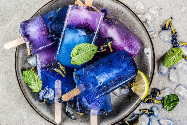 natürliche Bio-Sommerbonbons, hausgemachtes blaues und violettes Eis am Stiel mit Schmetterlingserbsenblumentee, grauer Betonhintergrund Kopierraum - Foto, Bild
