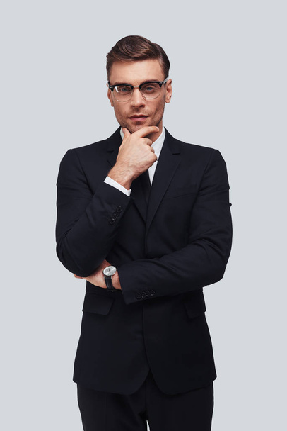 serious young man in full suit keeping hand on chin and looking at camera while standing against grey background - Фото, изображение