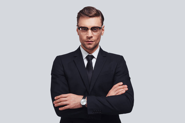 handsome young man in full suit keeping arms crossed while standing against grey background - Valokuva, kuva