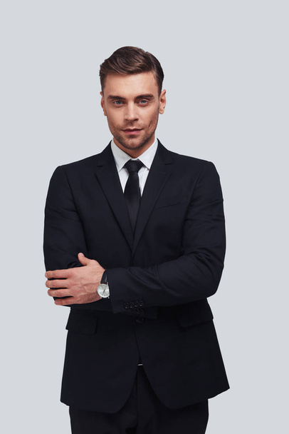 handsome young man in full suit keeping arms crossed while standing against grey background - Photo, Image