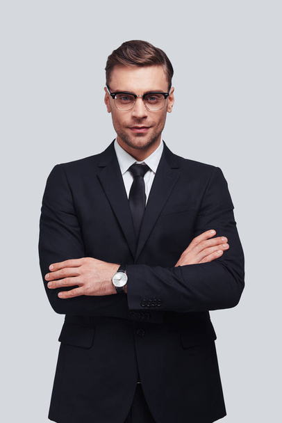 handsome young man in full suit keeping arms crossed while standing against grey background - Foto, Imagen