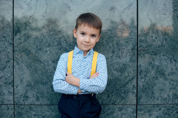 Close-up boy, future businessman, wearing yellow suspenders, dark blue pants and blue shirt against a green terracotta wall, with crossed arms and legs looking to camera, copy space. - Foto, Imagem