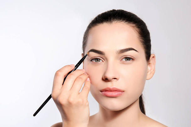 Young woman correcting shape of eyebrow with brush on light background - Fotó, kép