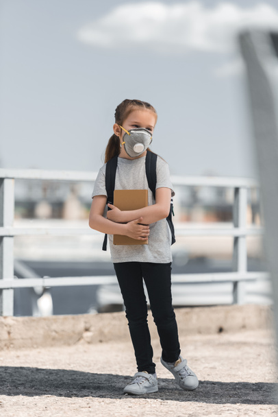 child in protective mask standing with bag and book on bridge, air pollution concept - 写真・画像