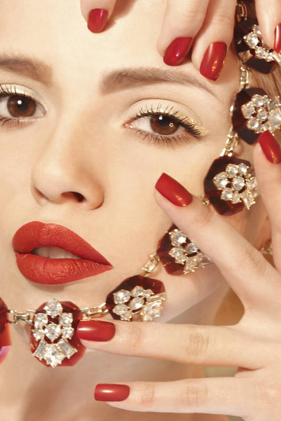 Manicura roja brillante en las uñas y lápiz labial rojo en el modelo de niña de cerca con un accesorio
. - Foto, imagen