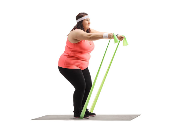Full length profile shot of an overweight woman exercising with a rubber band on a mat isolated on white background - Valokuva, kuva