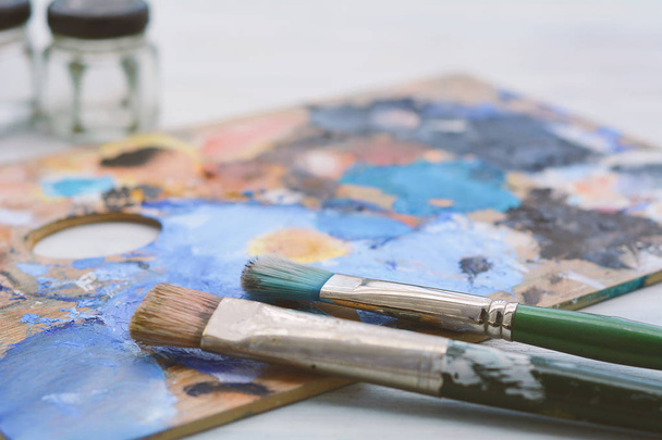 Artist's palette with colorful oil paint strokes, jars and paintbrushes on white wooden background. Art concept - 写真・画像