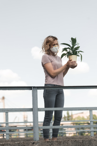 woman in protective mask holding green potted plant on bridge, air pollution concept - Zdjęcie, obraz