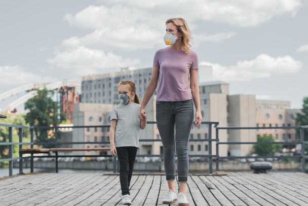 mother and daughter in protective masks holding hands on pier and looking away, air pollution concept - Photo, Image
