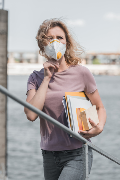 woman in protective mask standing on bridge with books and looking up, air pollution concept - Photo, Image