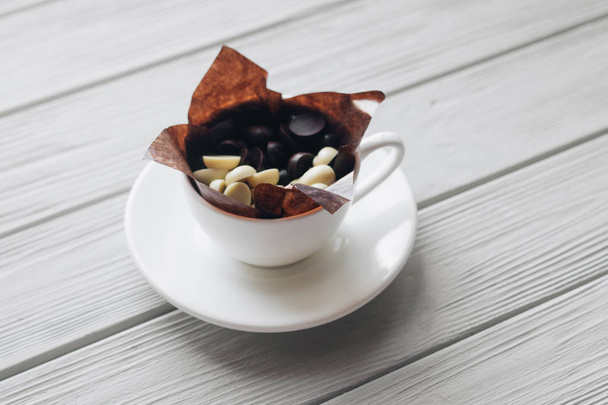 flat lay of delicious sweet chocolate drops in cup, white wooden background - Foto, Bild