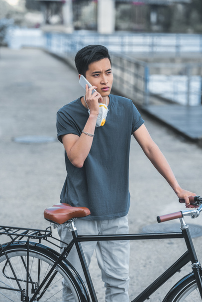 asian teen with protective mask talking by smartphone and leaning on bicycle, air pollution concept - Фото, изображение