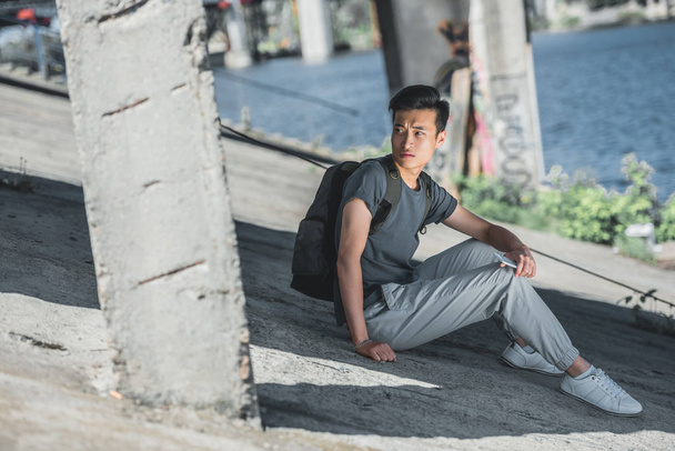 Asiatischer Teenager sitzt mit Smartphone unter Brücke in der Stadt und schaut weg - Foto, Bild