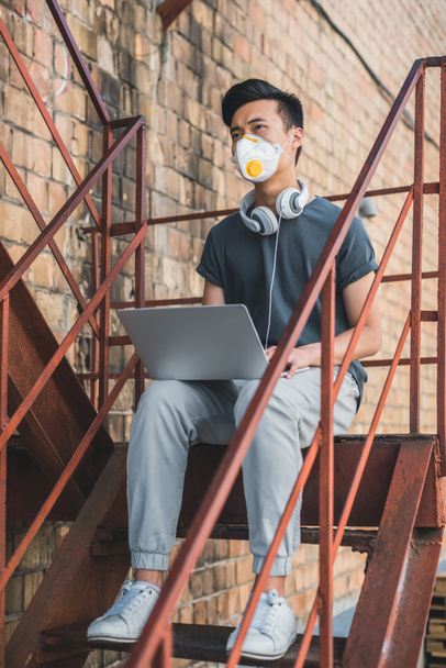 Aasialainen teini suojaava naamio istuu kannettavan tietokoneen kanssa portaissa ja katselee pois, ilmansaasteiden käsite
 - Valokuva, kuva