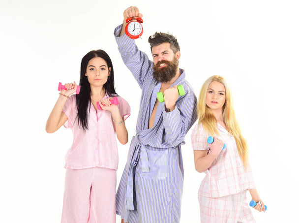 Fitness lifestyle concept. Trainer, macho with attractive girls do morning exercises. Cheerful friends with dumbbells in domestic clothes, isolated on white background. Bearded man holds alarm clock - Foto, Imagem