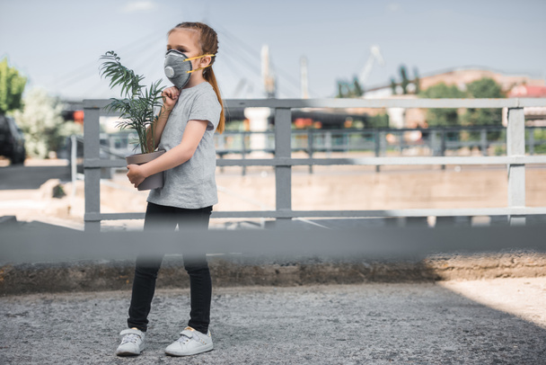 child in protective mask carrying green potted plant, air pollution concept - Foto, imagen