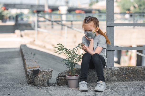 child in protective mask looking at green potted plant, air pollution concept - Photo, Image
