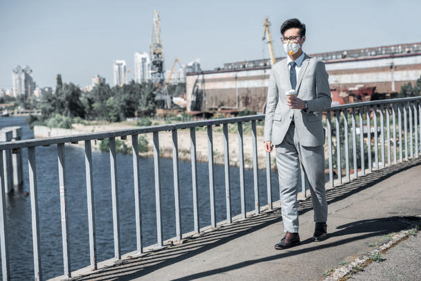 asian businessman in protective mask walking on bridge with coffee to go, air pollution concept - Фото, изображение