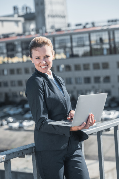 smiling attractive businesswoman standing with laptop on bridge and looking at camera - Φωτογραφία, εικόνα