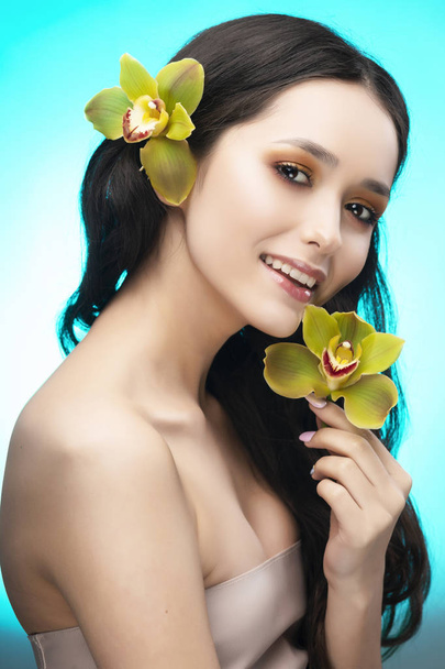 A beautiful brunette nude shoulders girl sensually holds a flower of an orchid in her hands. Nude make-up. Healthy smooth skin. Sky blue background. Copy space. Commercial and advertising design. - Foto, immagini