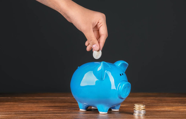 Woman putting coin into piggy bank on wooden table against dark background. Pension planning - Foto, Imagem
