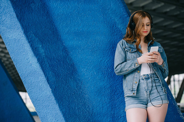 portrait of young fashionable woman in denim clothing and headphones using smartphone - Foto, Imagem