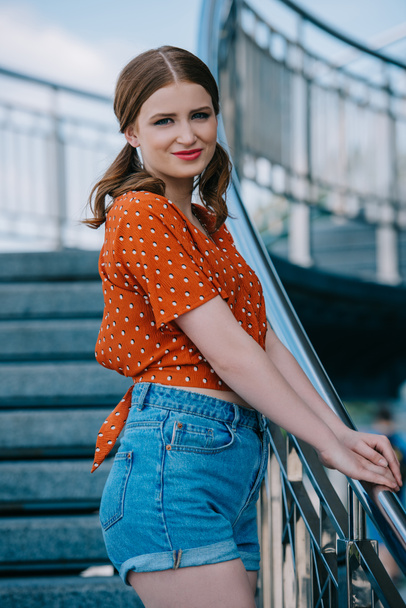 beautiful stylish girl in denim shorts standing on stairs and smiling at camera - Valokuva, kuva