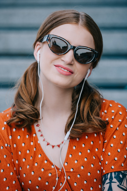 belle jeune femme en lunettes de soleil souriant à la caméra tout en écoutant de la musique dans les écouteurs sur la rue
 - Photo, image