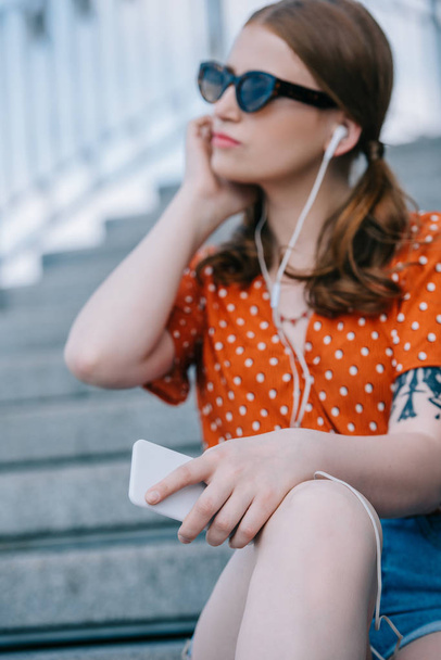 selective focus of girl in sunglasses and earphones listening music with smartphone while sitting on stairs - Foto, Imagen