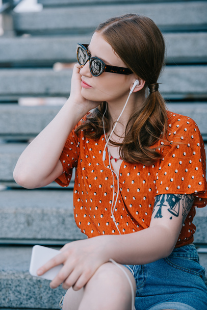 стильная девушка в солнечных очках и наушниках, слушающая музыку со смартфоном, сидя на лестнице
 - Фото, изображение