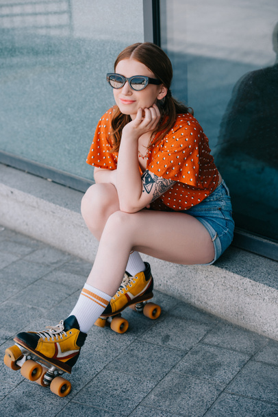 hoge hoekmening van stijlvolle lachende meisje in zonnebrillen en rolschaatsen zitten en weg op zoek op straat - Foto, afbeelding