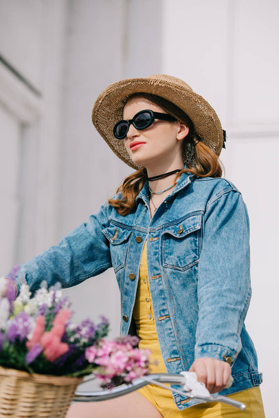 attractive girl in hat, sunglasses and denim jacket riding bicycle on street  - Foto, imagen